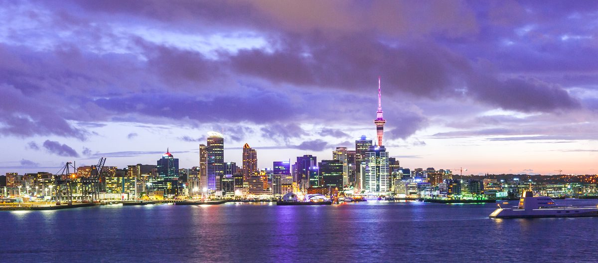 The Auckland city skyline and Sky Tower lit up at dusk across the harbour, under purple cloud