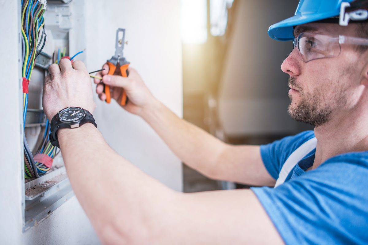 An electrician in a blue hard hat and safety glasses stripping a wire with pliers at a wall box full of coloured cables