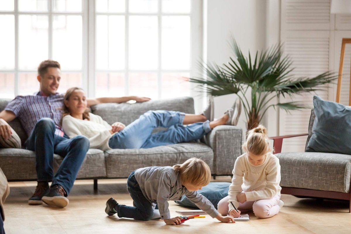 Two adults relaxing on a grey sofa while two children draw on the floor of a bright lounge with tall windows