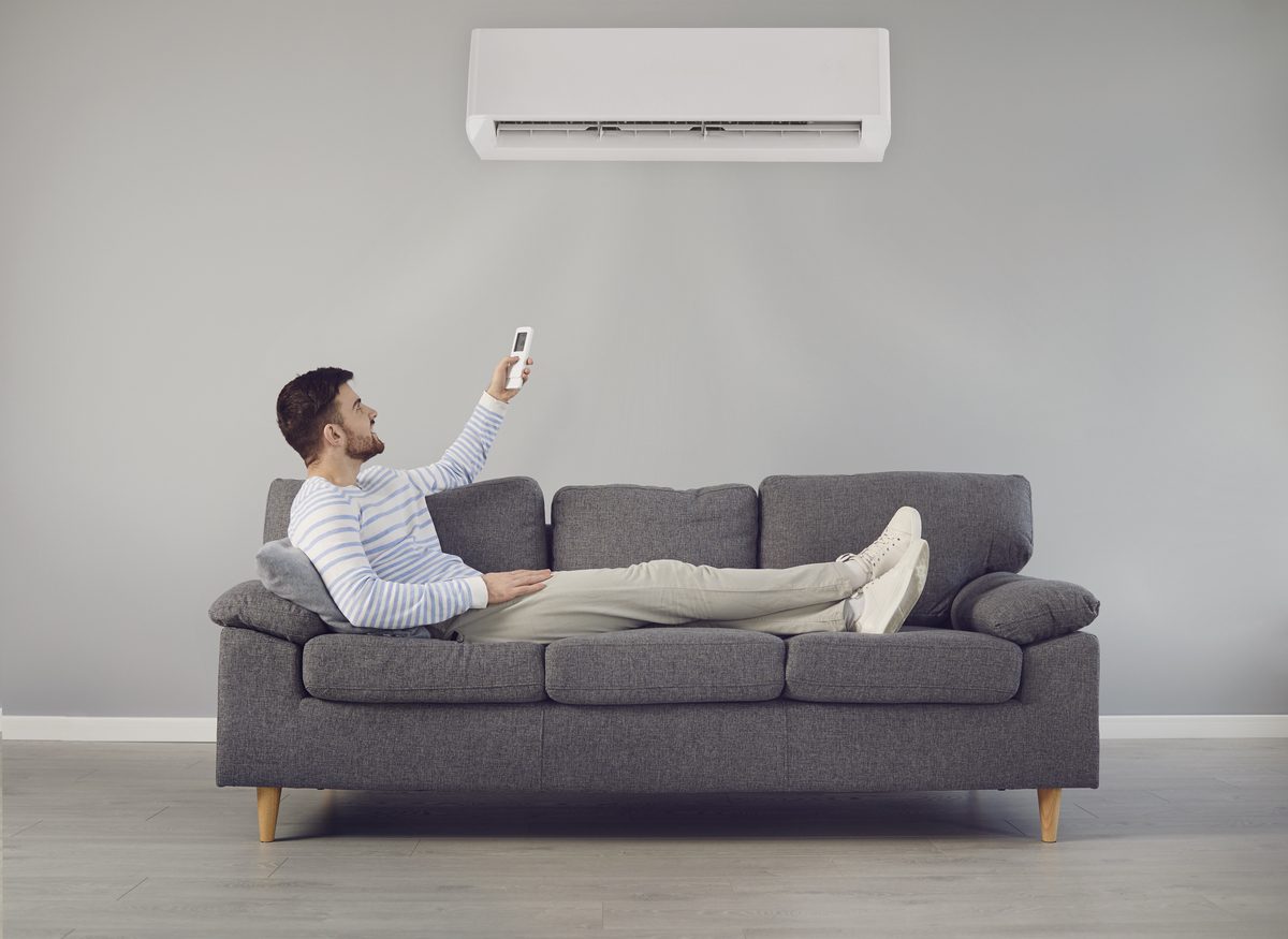 A man lying on a grey sofa pointing a remote control at a white heat pump mounted high on the wall